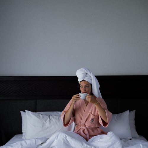 Personne assise sur un lit d’hôtel à Amber Kampot, serviette sur la tête, buvant un café avec un petit-déjeuner en chambre.