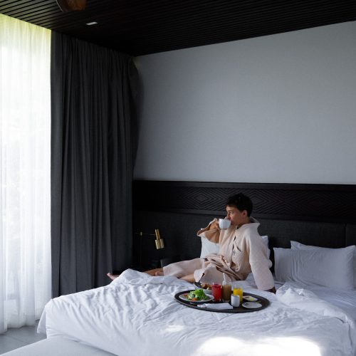 Personne assise sur un lit d’hôtel à Amber Kampot, dégustant un café avec un petit-déjeuner servi en chambre.