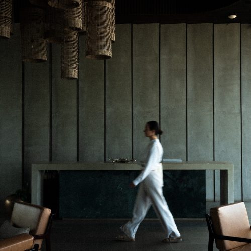 Personne marchant dans le lobby de l’hôtel Amber Kampot, sous des suspensions tressées et un design épuré.