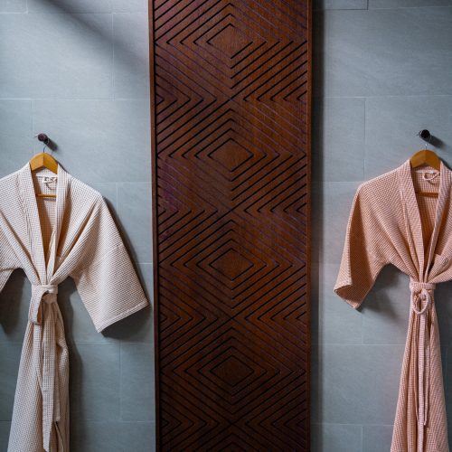Deux peignoirs suspendus dans la salle de bain design de l’hôtel Amber Kampot, avec panneau mural en bois sculpté.