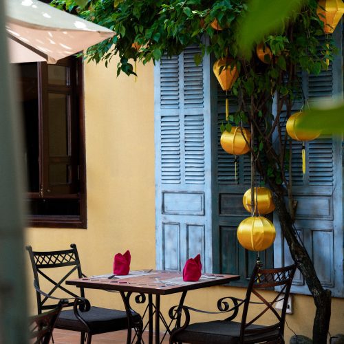 Table de restaurant en terrasse à La Siesta Hoi An, entourée de lanternes jaunes, de verdure et de volets bleus.