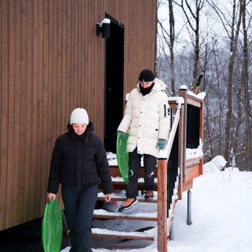 Deux personnes sortant d’un chalet du domaine Le Vertendre en hiver, tenant des luges dans la neige.