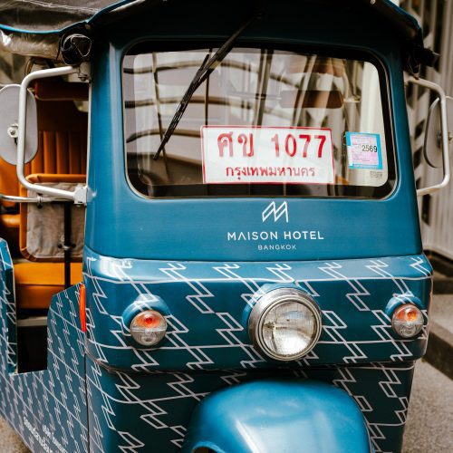 Tuk-tuk bleu personnalisé aux couleurs du Maison Hotel Bangkok, stationné devant l’hôtel.
