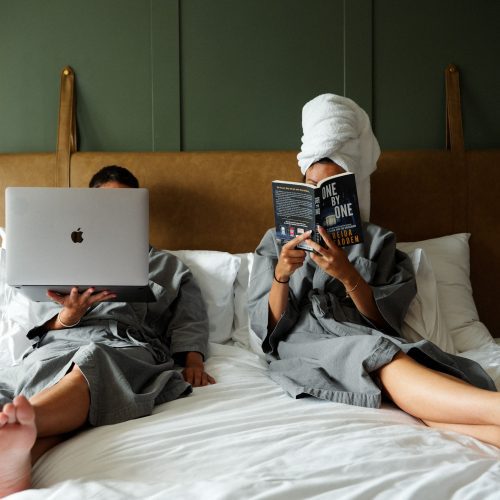 Deux voyageurs se détendent sur un lit king-size dans la PHANA Suite King du Maison Hotel Bangkok, entre lecture et travail.