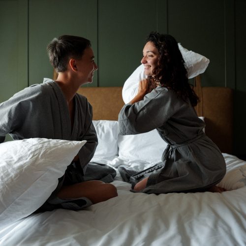 Deux personnes en peignoir se font face sur le lit et s’apprêtent à se lancer un oreiller, dans la Phana Suite King au Maison Hotel Bangkok.