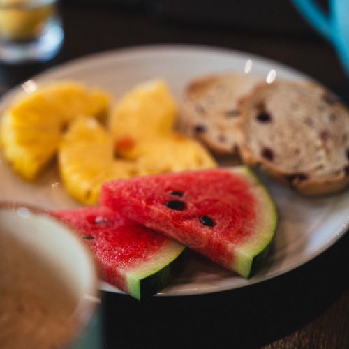 Assiette de petit-déjeuner avec pastèque, ananas et tranches de pain, avec une tasse au premier plan, au Maison Hotel Bangkok.