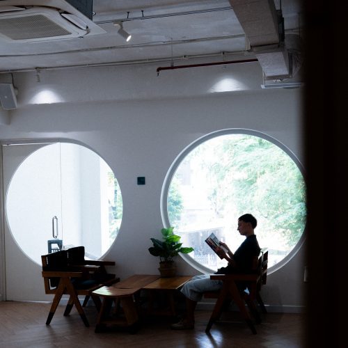 Personne assise lisant un livre dans l’espace coworking de Meander Saigon, éclairé par deux grandes fenêtres circulaires.
