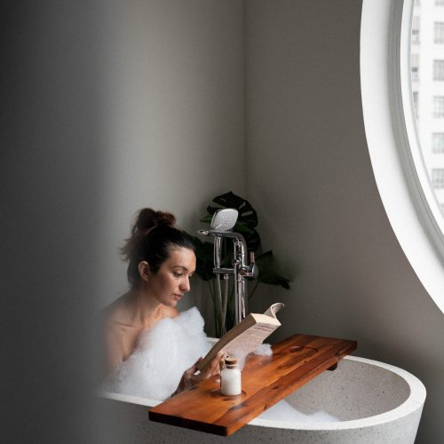 Personne lisant un livre dans une baignoire de la suite de l’hôtel Meander Saigon, près d’une grande fenêtre circulaire.