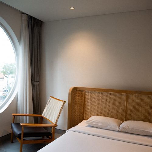 Lit double et fauteuil près d’une fenêtre circulaire dans une chambre de l’hôtel Meander Saigon.