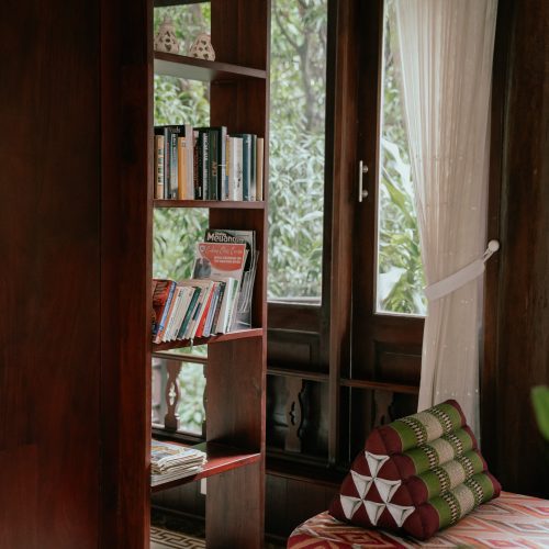Étagère en bois avec livres et magazines, à côté d’une banquette et d’une fenêtre donnant sur la végétation, au Parasol Blanc à Luang Prabang.