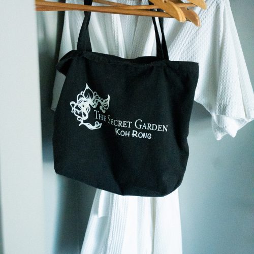 Sac tote bag noir « The Secret Garden Koh Rong » suspendu à des cintres devant un peignoir blanc dans une penderie.
