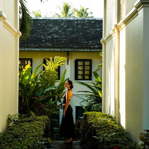 Personne debout dans une allée verdoyante du Villa Maly à Luang Prabang, entre bâtiments coloniaux et végétation tropicale.