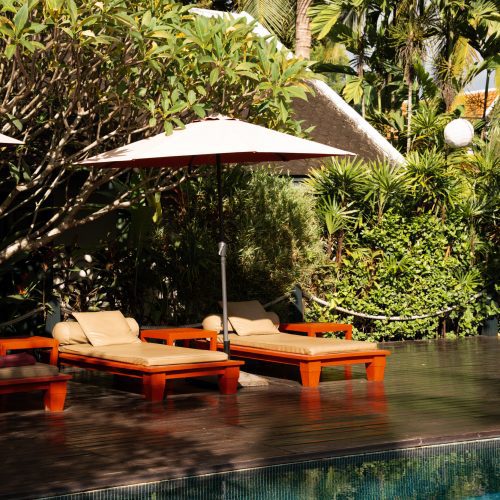 Transats en bois et coussins beiges au bord de la piscine du Villa Maly, entourés d’une végétation tropicale luxuriante à Luang Prabang.