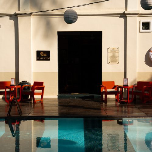 Façade du bar du Villa Maly à Luang Prabang avec chaises rouges sur la terrasse en bois, reflétées dans la piscine au premier plan.