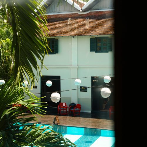 Piscine du Villa Maly à Luang Prabang vue à travers des feuilles tropicales, avec lanternes suspendues et bâtiment colonial en arrière-plan.