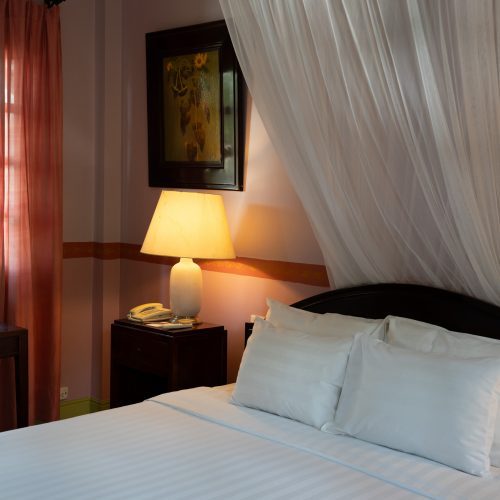 Lit à baldaquin dans une chambre du Villa Maly à Luang Prabang, avec moustiquaire blanche, lampe allumée et décoration chaleureuse.