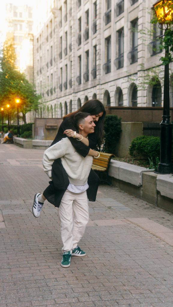 Couple de femmes souriantes, l’une portant l’autre sur son dos dans une rue urbaine.